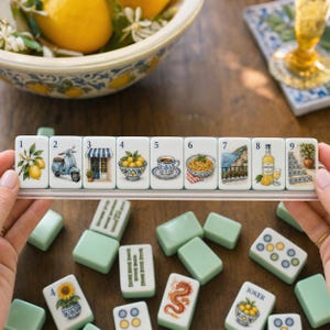May include: A set of nine rectangular white tiles arranged in a clear plastic rack, each featuring a unique illustration including lemons, a scooter, a storefront, a bowl of fruit, a coffee cup, pasta, a coastal view, limoncello, and stairs.