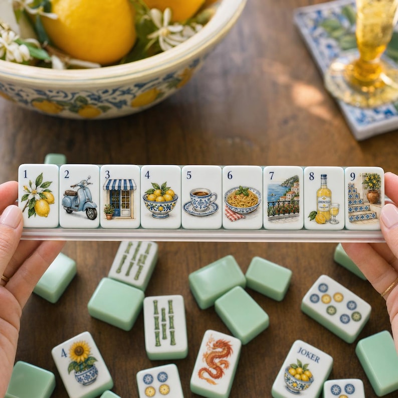 May include: A set of nine rectangular white tiles arranged in a clear plastic rack, each featuring a unique illustration including lemons, a scooter, a storefront, a bowl of fruit, a coffee cup, pasta, a coastal view, limoncello, and stairs.