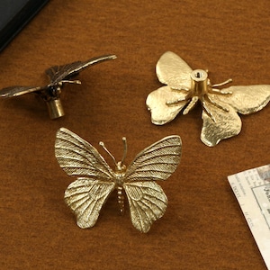 May include: Three gold butterfly-shaped drawer pulls. Two of the pulls have a more realistic butterfly design, while the third pull has a more stylized design.