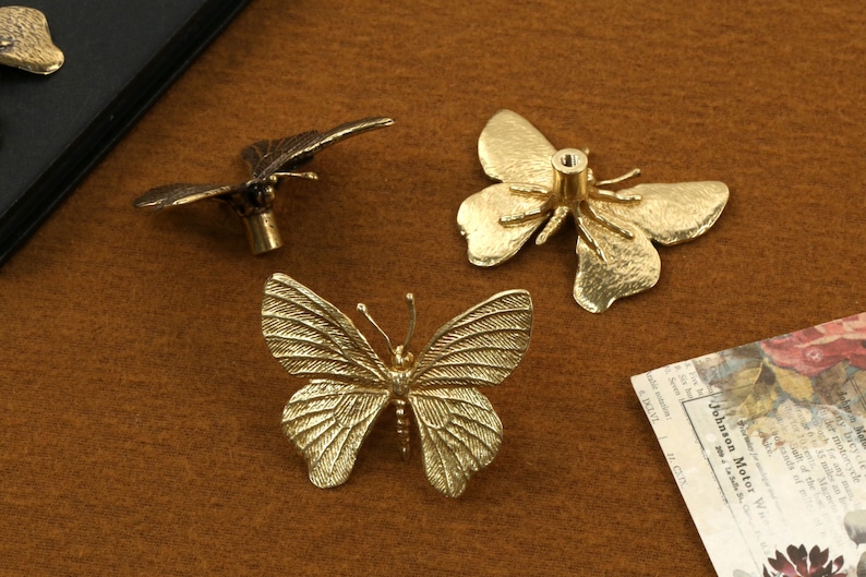 May include: Three gold butterfly-shaped drawer pulls. Two of the pulls have a more realistic butterfly design, while the third pull has a more stylized design.