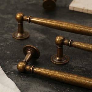 Pu&ograve; includere: Primo piano di diverse maniglie per mobili in ottone antico. Le maniglie hanno un'impugnatura cilindrica con un'estremit&agrave; arrotondata e una base circolare. La finitura &egrave; un colore ottone screziato e invecchiato, che suggerisce un'estetica vintage. Le maniglie sono disposte su una superficie scura e strutturata.