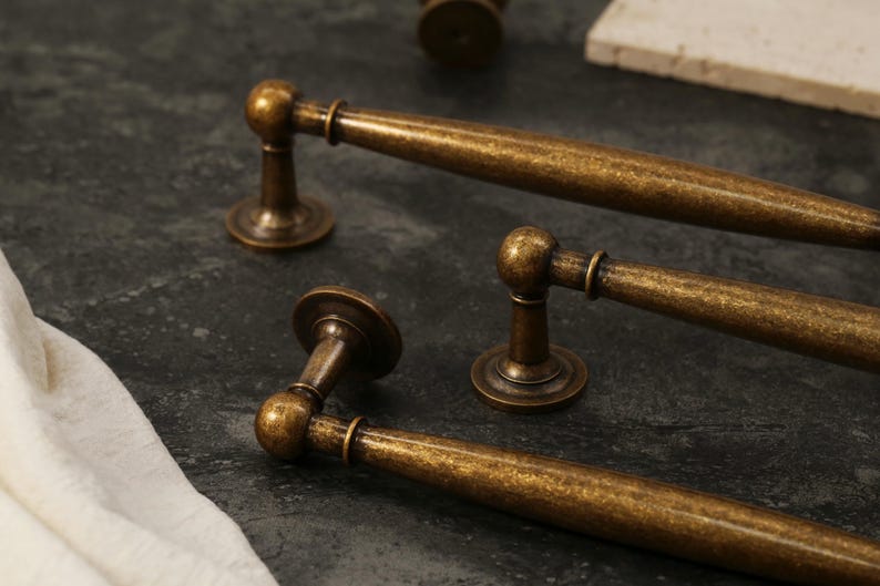 Pu&ograve; includere: Primo piano di diverse maniglie per mobili in ottone antico. Le maniglie hanno un'impugnatura cilindrica con un'estremit&agrave; arrotondata e una base circolare. La finitura &egrave; un colore ottone screziato e invecchiato, che suggerisce un'estetica vintage. Le maniglie sono disposte su una superficie scura e strutturata.