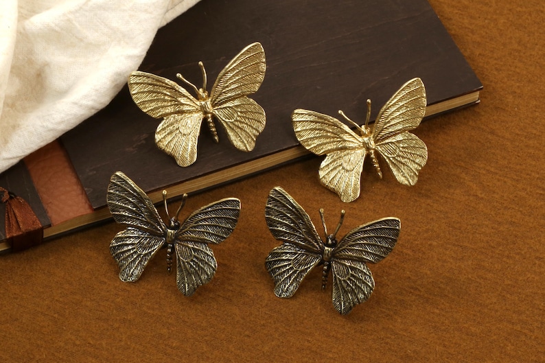 May include: Four decorative metal butterfly ornaments in gold and bronze finishes. The butterflies are arranged on a brown surface.