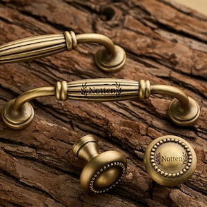 May include: Antique brass cabinet hardware set, including two handles and a knob. The handles feature a ribbed design with curved ends. The knob and a round pull have the word "Notten" and a laurel wreath design.