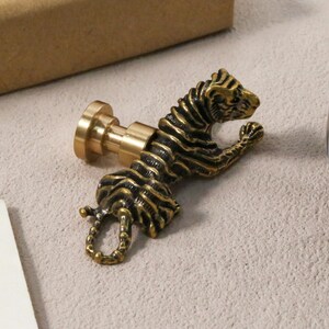 K&ouml;nnte beinhalten: Ein detaillierter Wachssiegelstempel in Form eines Tigers aus Messing. Der Tiger ist in einer dynamischen Pose mit detaillierten Streifen dargestellt. Der Stempel hat eine runde Basis und einen zylindrischen Griff. Das Design ist antik und dekorativ.