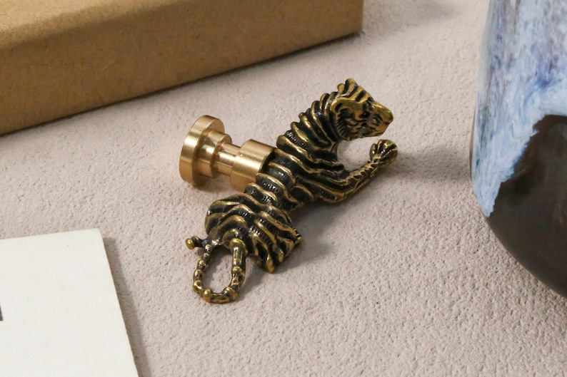 K&ouml;nnte beinhalten: Ein detaillierter Wachssiegelstempel in Form eines Tigers aus Messing. Der Tiger ist in einer dynamischen Pose mit detaillierten Streifen dargestellt. Der Stempel hat eine runde Basis und einen zylindrischen Griff. Das Design ist antik und dekorativ.