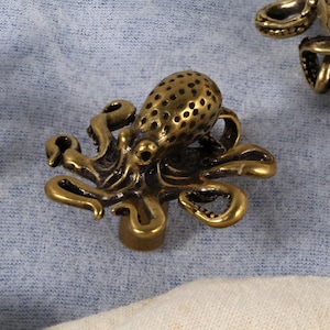 May include: A detailed, antique brass octopus-shaped cabinet knob. The knob features a textured body with small holes and curled tentacles. The octopus is centered on a light blue fabric background.