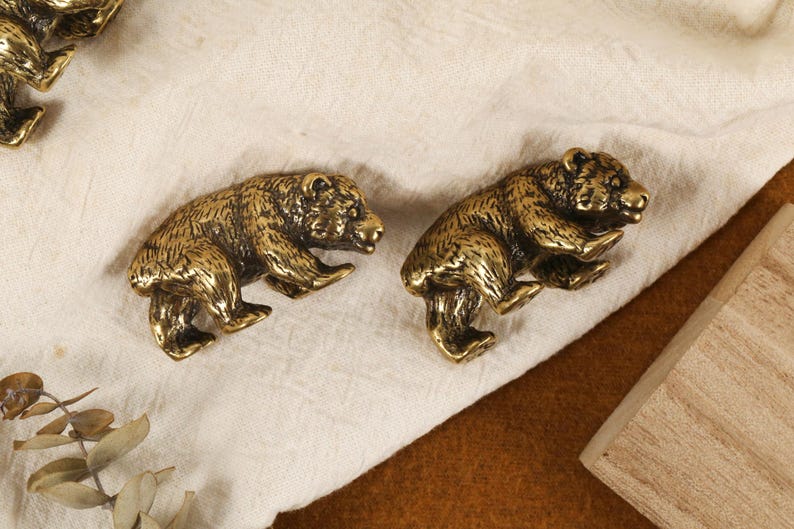 Puede incluir: Dos figuras de oso de lat&oacute;n antiguo, cada una en posici&oacute;n de caminar, con textura de pelaje detallada. Los osos est&aacute;n colocados sobre una tela color crema, con un bloque de madera visible en el fondo. La est&eacute;tica general es r&uacute;stica y decorativa.