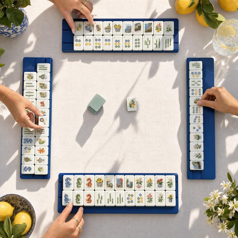 May include: A Mahjong game set featuring four blue tile racks arranged in a square on a white tablecloth. Each rack holds white tiles decorated with colorful botanical and dragon illustrations. A single loose tile sits in the center.