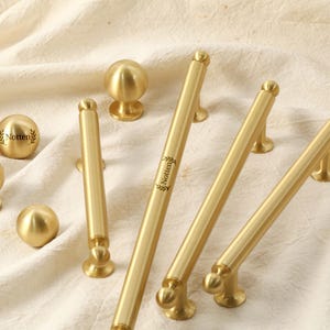 May include: Various brass cabinet handles and knobs displayed on a cream-coloured cloth. The hardware features a polished finish and classic design. The brand name "Notten" is visible on some pieces.