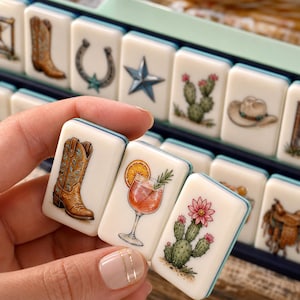 May include: A set of rectangular mahjong tiles featuring colorful illustrations of western-themed icons including cowboy boots, horseshoes, stars, cacti, hats, and a cocktail glass with an orange slice and a sprig of rosemary.