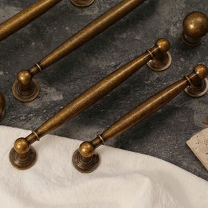 Pu&ograve; includere: Una collezione di ferramenta per mobili in ottone antico, tra cui diverse maniglie e pomelli rotondi. L'hardware ha una finitura calda e invecchiata ed &egrave; disposto su una superficie scura e strutturata con un panno bianco.