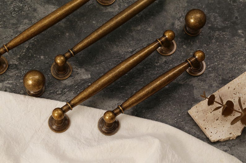 Pu&ograve; includere: Una collezione di ferramenta per mobili in ottone antico, tra cui diverse maniglie e pomelli rotondi. L'hardware ha una finitura calda e invecchiata ed &egrave; disposto su una superficie scura e strutturata con un panno bianco.