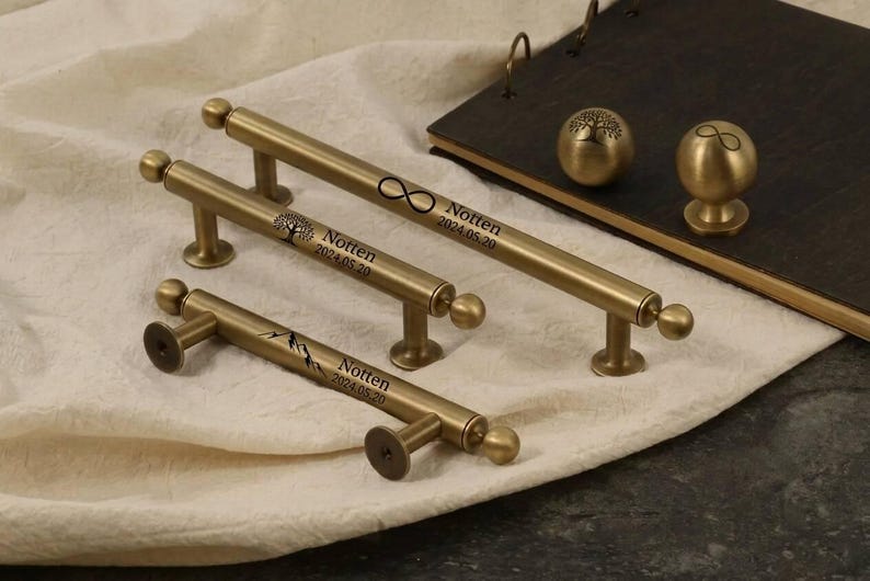May include: A collection of brass-toned cabinet hardware, including handles and knobs. The handles feature engraved designs and the text "Notten 2024.05.20". The knobs have tree and infinity symbols. The hardware is displayed on a white cloth.