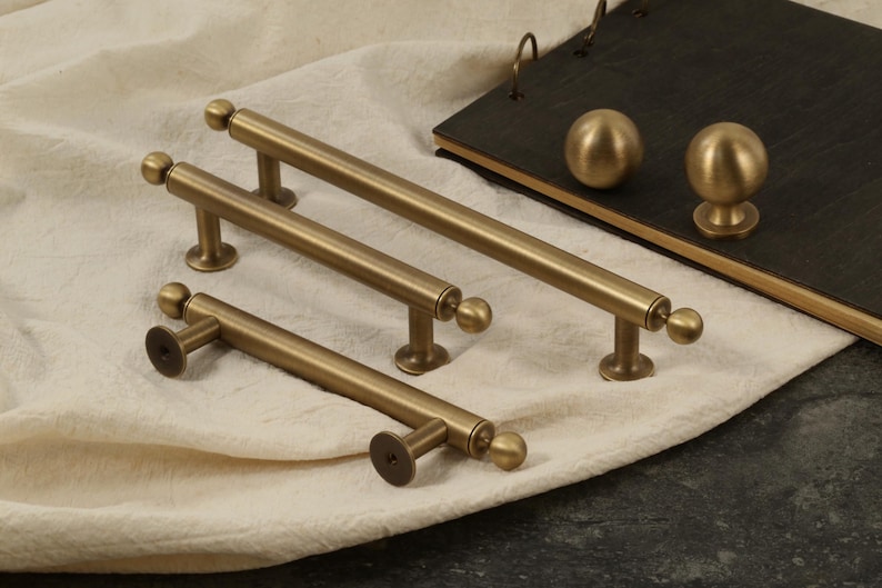 K&ouml;nnte beinhalten: Eine Sammlung antiker Messing-Schrankbeschl&auml;ge, darunter Stangengriffe und runde Kn&ouml;pfe. Die Griffe variieren in der Gr&ouml;&szlig;e, mit einem zylindrischen Stab und Kugelabschl&uuml;ssen. Die Kn&ouml;pfe sind kugelf&ouml;rmig.