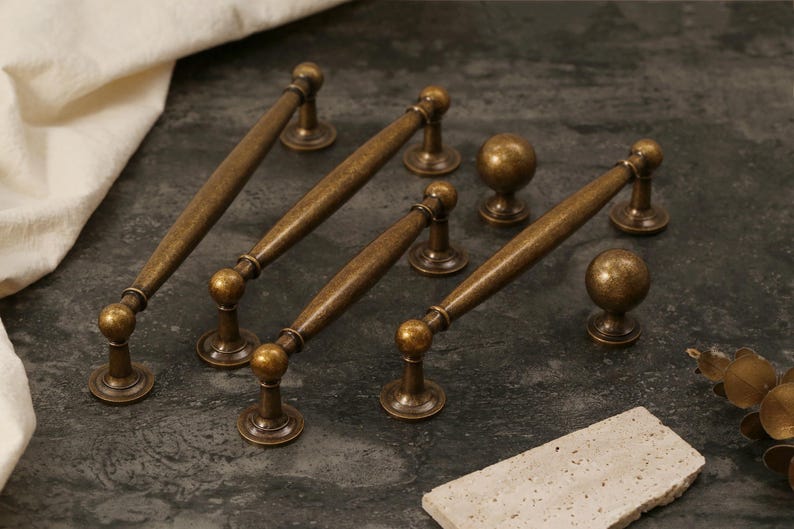 Pu&ograve; includere: Una collezione di ferramenta per mobili in ottone antico, tra cui quattro maniglie e due pomelli sferici. La ferramenta ha una finitura calda e invecchiata ed &egrave; disposta su una superficie scura e strutturata. Le maniglie misurano circa 15 cm.