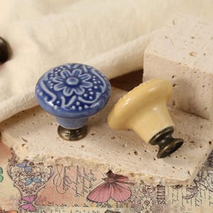 Op de afbeelding: Drie keramische ladeknopjes met bloemmotieven. De knoppen zijn in tinten roze, blauw en cr&egrave;me. De knoppen worden getoond op een getextureerd oppervlak.