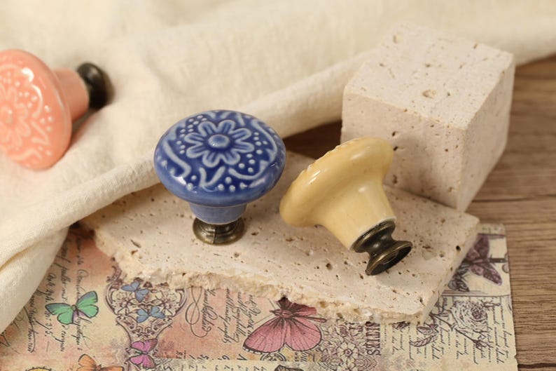 Op de afbeelding: Drie keramische ladeknopjes met bloemmotieven. De knoppen zijn in tinten roze, blauw en cr&egrave;me. De knoppen worden getoond op een getextureerd oppervlak.