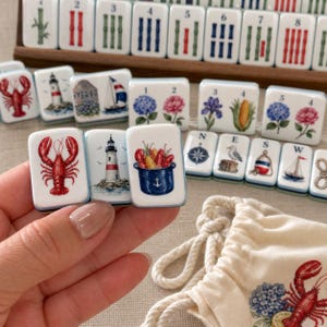 May include: A set of white rectangular mahjong tiles featuring colorful illustrations of lobsters, lighthouses, sailboats, flowers, and nautical symbols. Some tiles display numbers and letters. A matching cream-colored drawstring bag is included.