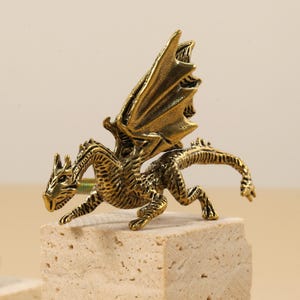 May include: A detailed, antique gold-colored dragon figurine with outstretched wings. The dragon is posed on a textured, light-colored block. The figurine has intricate detailing and a mythical theme.