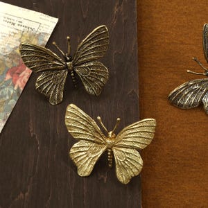 May include: Three gold-toned metal butterfly ornaments. The butterflies are arranged on a brown wooden surface and a brown fabric surface.