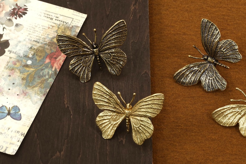 May include: Three gold-toned metal butterfly ornaments. The butterflies are arranged on a brown wooden surface and a brown fabric surface.