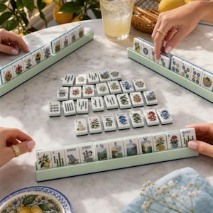 May include: A Mahjong game set featuring white tiles with colorful illustrations of flowers, dragons, and scenery, arranged in light green racks and on a marble surface. The tiles display various numbers, letters, and the word JOKER.