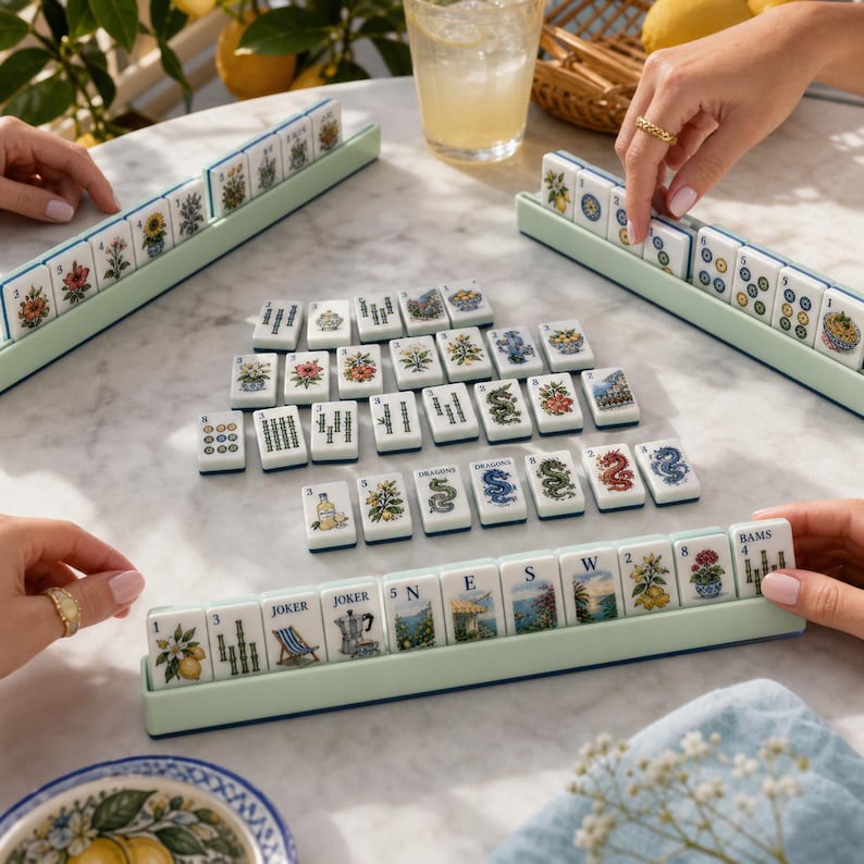 May include: A Mahjong game set featuring white tiles with colorful illustrations of flowers, dragons, and scenery, arranged in light green racks and on a marble surface. The tiles display various numbers, letters, and the word JOKER.