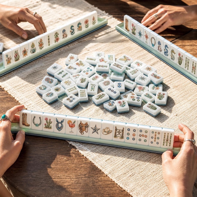 May include: A Mahjong game set featuring white tiles with colorful illustrations of western-themed icons like cowboy boots, cacti, horseshoes, and stars, arranged in racks and a central pile on a textured woven surface.