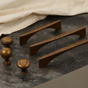 May include: A collection of antique bronze cabinet hardware, including two styles of handles and two styles of knobs. The handles have a curved design, and the knobs are round. The hardware is arranged on a dark gray surface.