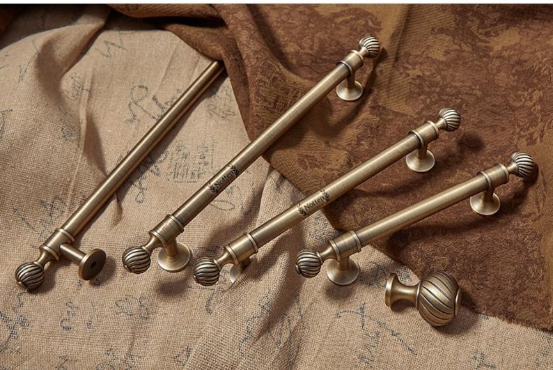 May include: A selection of antique brass cabinet pulls and handles. The hardware features a ribbed ball design and a round base. The pulls are arranged on a textured, neutral-toned fabric.