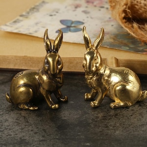 May include: Two brass rabbit figurines. The rabbits are in a sitting position with their ears upright. The figurines have a shiny, golden finish. They are placed on a dark surface with a blurred background.