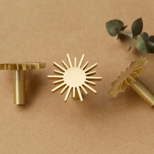 May include: Three gold metal cabinet knobs with a sunburst design. The knobs have a round base and a long, thin stem.