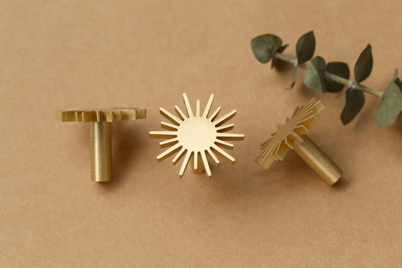 May include: Three gold metal cabinet knobs with a sunburst design. The knobs have a round base and a long, thin stem.