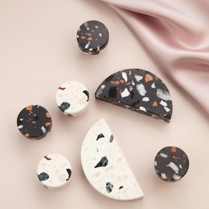 Könnte beinhalten: Eine Auswahl an dekorativen Schrankknöpfen und -griffen aus Terrazzo. Die Knöpfe und Griffe weisen ein gesprenkeltes Design mit schwarzen, weißen, orangefarbenen und grünen Fragmenten auf. Die Formen umfassen Kreise und Halbkreise. Der Hintergrund ist hellrosa.