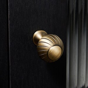 May include: A close-up shot of a brass cabinet knob with a spherical design and grooved detailing. The knob is mounted on a black wooden cabinet door. The hardware has a vintage aesthetic.