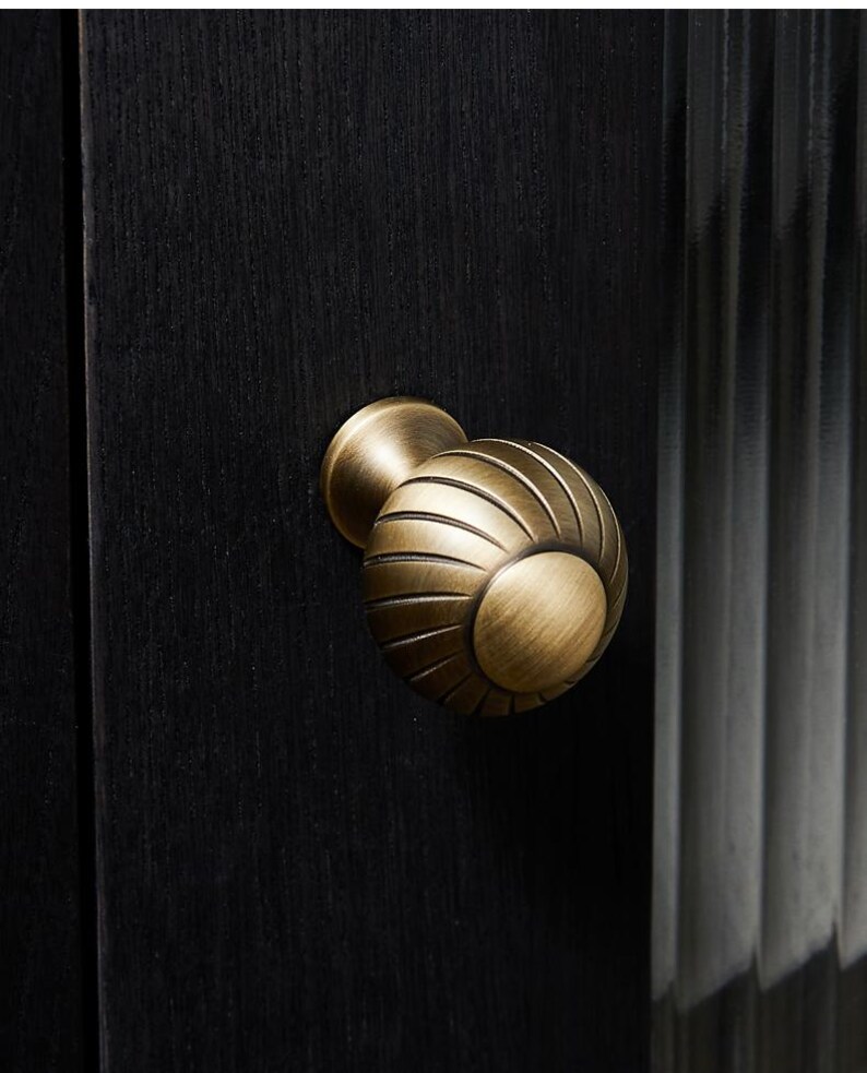 May include: A close-up shot of a brass cabinet knob with a spherical design and grooved detailing. The knob is mounted on a black wooden cabinet door. The hardware has a vintage aesthetic.