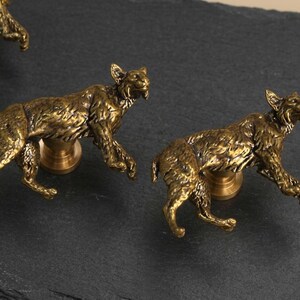 May include: A set of three brass-toned cabinet knobs, each shaped like a crouching lynx with detailed fur texture. The metallic figures are mounted on circular bases, designed to function as hardware pulls for furniture or cabinetry doors.