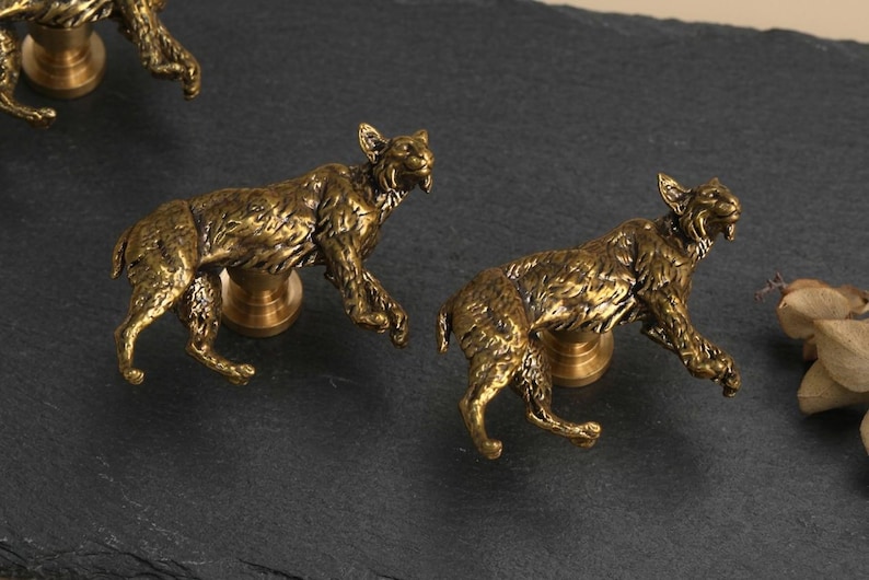 May include: A set of three brass-toned cabinet knobs, each shaped like a crouching lynx with detailed fur texture. The metallic figures are mounted on circular bases, designed to function as hardware pulls for furniture or cabinetry doors.