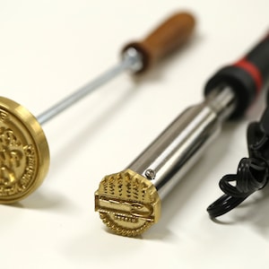May include: Two metal branding irons with wooden handles. One iron has a round head with the text "The Original " and the other has a rectangular head with a tree design.