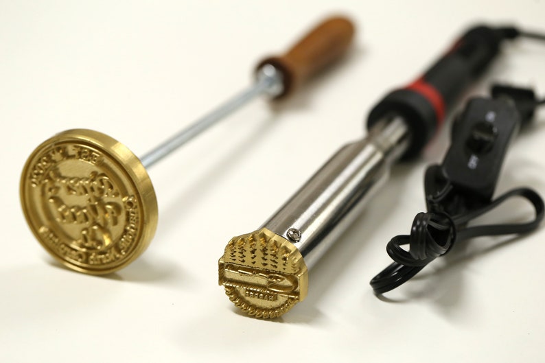 May include: Two metal branding irons with wooden handles. One iron has a round head with the text "The Original " and the other has a rectangular head with a tree design.