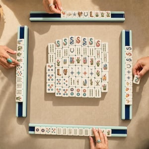 May include: A Mahjong game set featuring white tiles with colorful illustrations of cacti, cowboy boots, horseshoes, stars, and cocktails, arranged in a central square formation with four light blue tile racks.