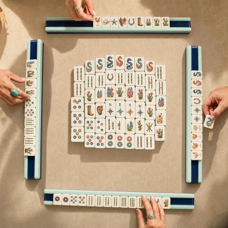 May include: A Mahjong game set featuring white tiles with colorful illustrations of cacti, cowboy boots, horseshoes, stars, and cocktails, arranged in a central square formation with four light blue tile racks.