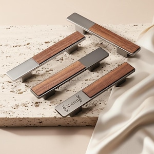 May include: Four cabinet pulls with a modern design. Each pull features a rectangular, brushed metal end and a rectangular, dark wood center. The pulls are arranged on a textured, light-colored surface. One pull has the word "Notten" and a leaf design.