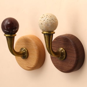 May include: Two wall-mounted hooks featuring circular wooden bases, one light-toned and one dark-toned. Each hook has a curved brass-colored metal arm topped with a spherical finial, one dark brown and polished, the other beige and textured.