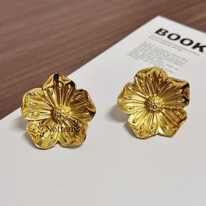 以下が含まれることがあります： 金色の花の形をしたスタッドピアス。ピアスは、詳細な花びらのデザインとテクスチャード加工された中央部を備えています。「Notteny」の文字が片方のピアスに見えます。ピアスは明るい色の表面に表示されています。