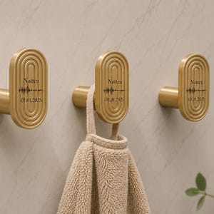 Pode incluir: Três ganchos de parede dourados com um design oval e uma base cilíndrica. Cada gancho tem o nome "Notten" e a data "01.01.2025" impressos. Uma toalha bege está pendurada num dos ganchos.