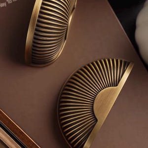 May include: Two antique brass cabinet pulls with a half-circle, sunburst design. The pulls have a ribbed texture and a warm, aged finish, ideal for adding a vintage touch to furniture.