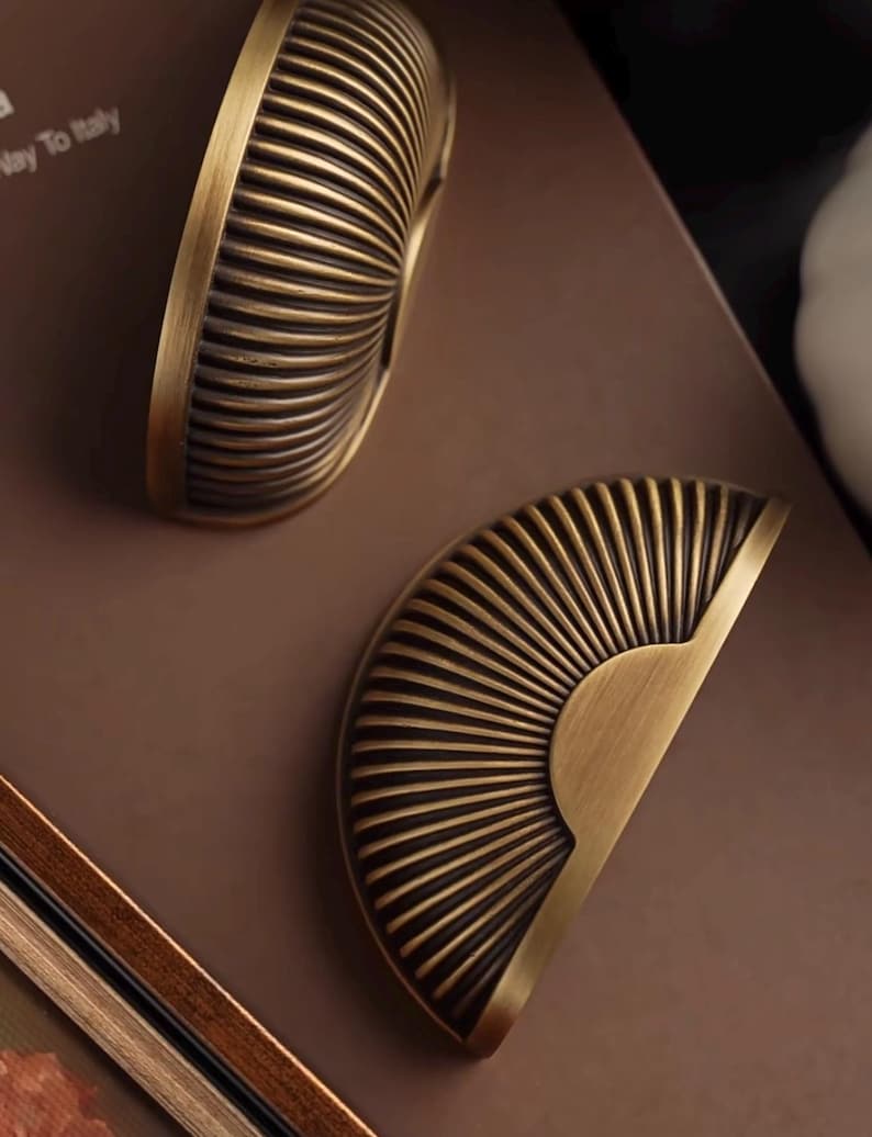 May include: Two antique brass cabinet pulls with a half-circle, sunburst design. The pulls have a ribbed texture and a warm, aged finish, ideal for adding a vintage touch to furniture.