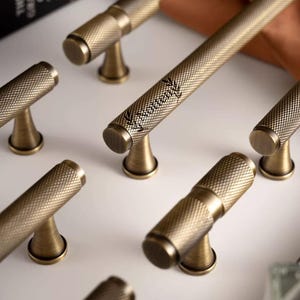 Könnte beinhalten: Antike Messing-Schrankgriffe mit strukturiertem Rautenmuster. Ein langer Griff trägt das Wort "Vintrent" in Schreibschrift. Die Griffe haben eine zylindrische Form mit rundem Sockel. Ideal für die Möbelrestaurierung.